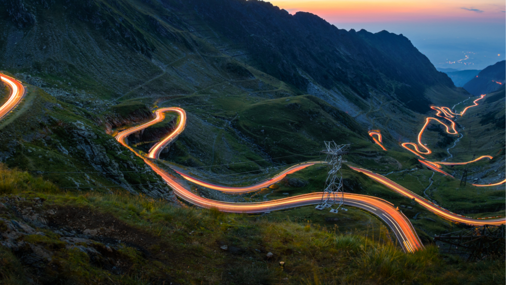 Une route de montagne sinueuse (Transfăgărășan) au crépuscule, transformée en hub énergétique mobile Revolt Power Green avec des panneaux solaires, des éoliennes, des unités de stockage Northvolt et des bornes de recharge pour véhicules électriques.